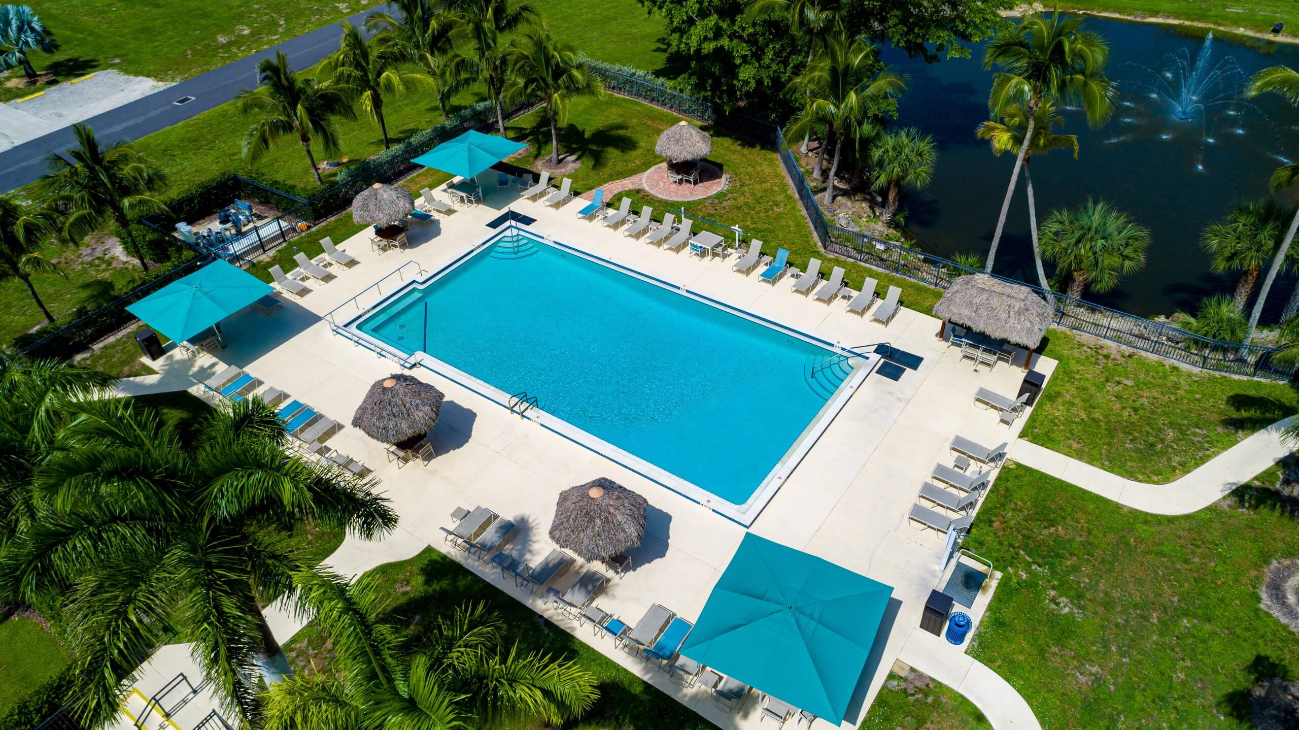 Aerial view of a rectangular swimming pool with lounge chairs, umbrellas, and a nearby pond with a fountain in Bonita Springs, FL