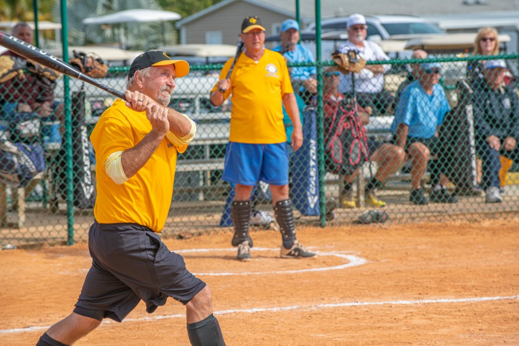 Older man swinging a bat during a softball game, with teammates and spectators watching from behind the fence in Bonita Springs, FL