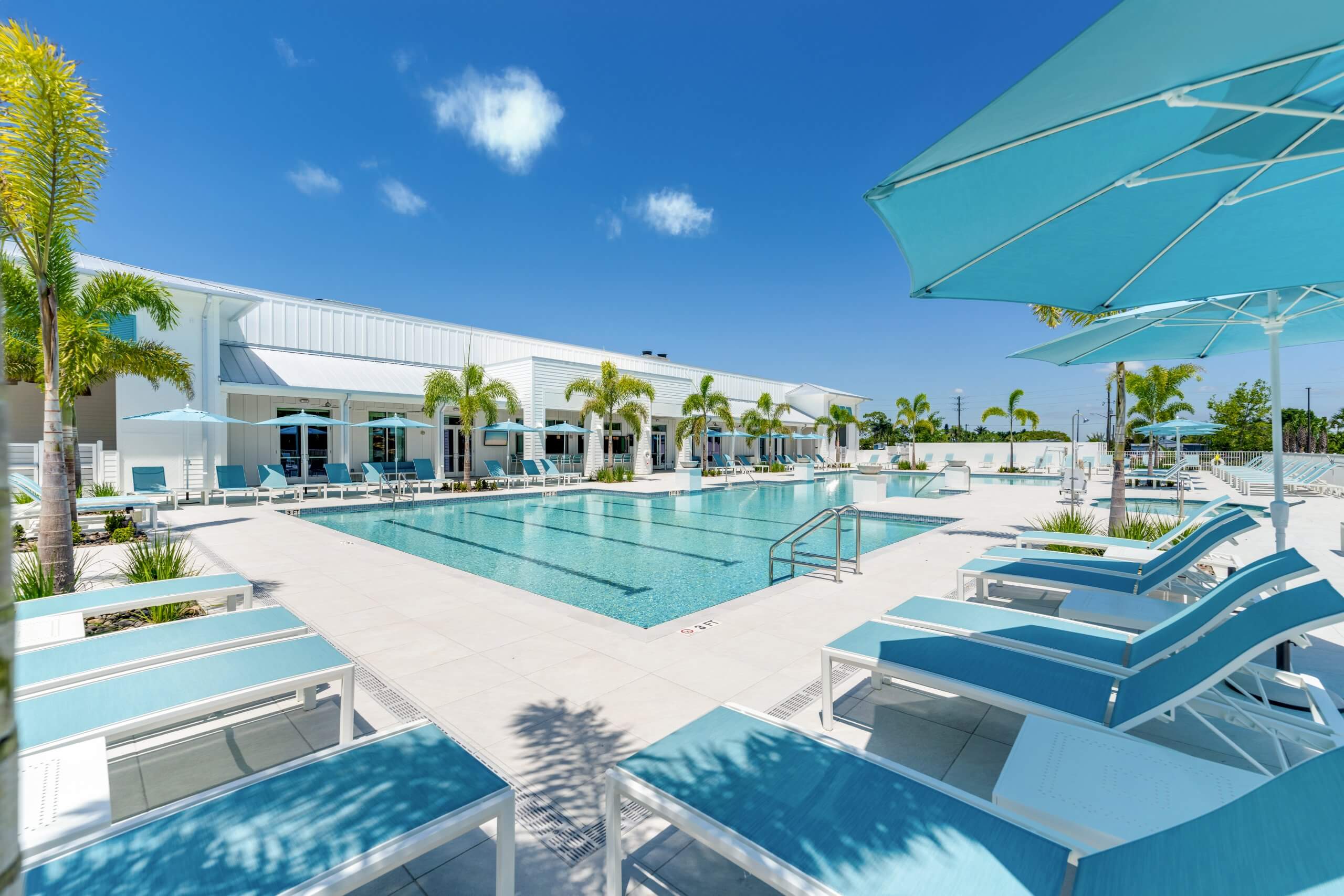 Resort-style swimming pool with turquoise lounge chairs and umbrellas, next to a modern white clubhouse in Bonita Springs, FL