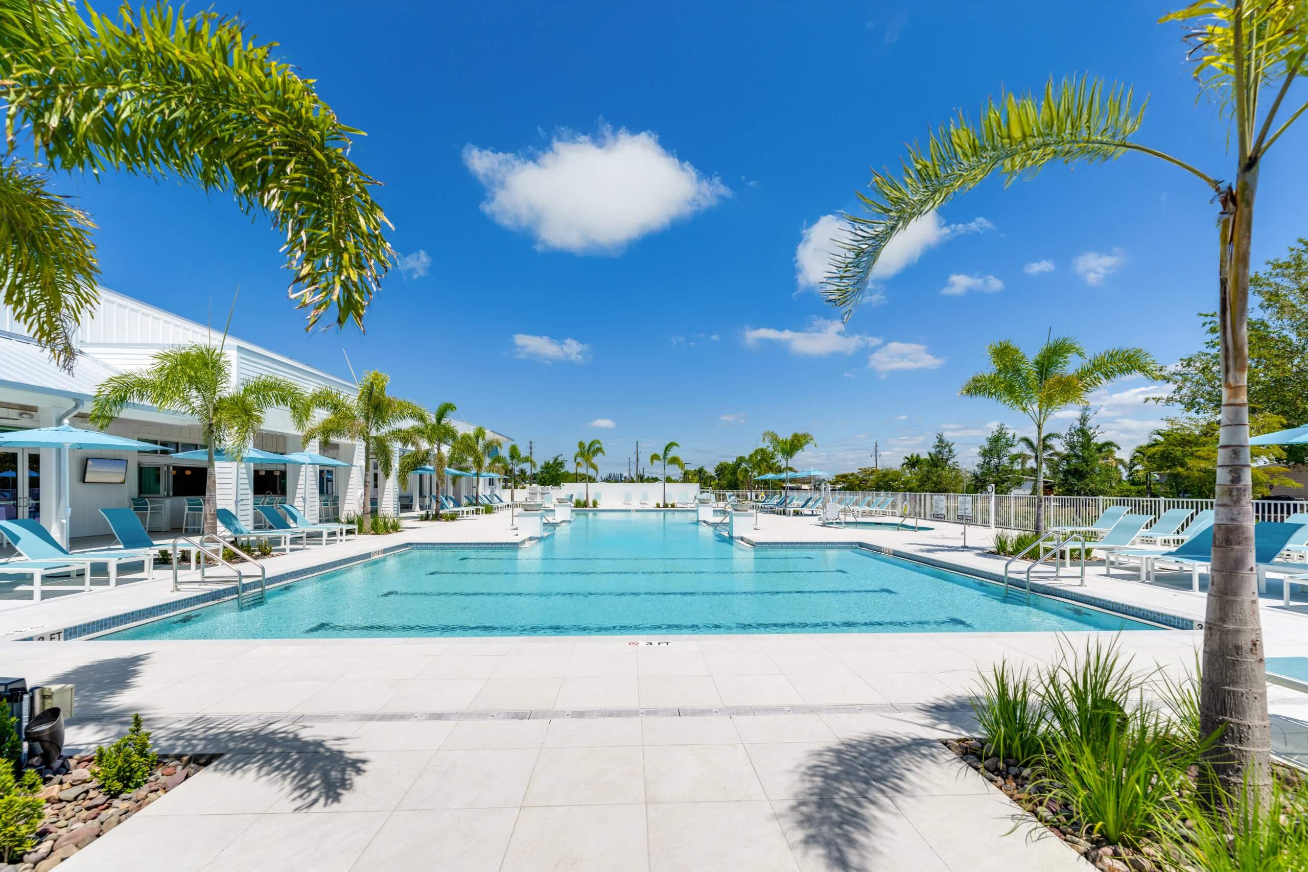Resort-style swimming pool with palm trees, lounge chairs, and blue umbrellas under a clear blue sky in Bonita Springs, FL